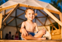 Smiling boy sits barefoot cross-legged in lotus position on wooden floor and looks up at the morning sun.