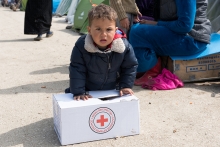 Worried young boy in refugee camp leans on Red Cross relief box.